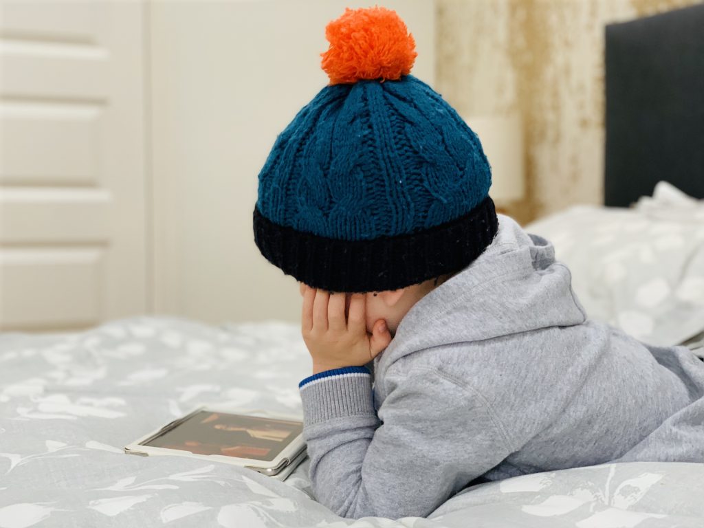 A toddler wearing a wooly hat watches an ipad lying on a bed