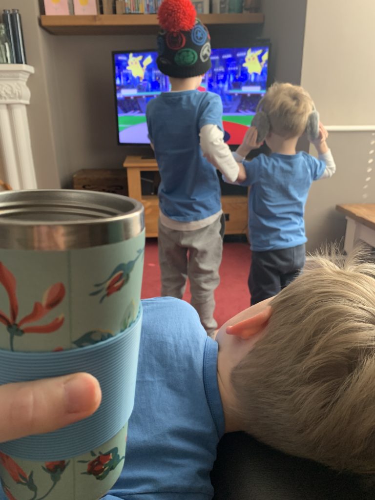 Toddler lies on mummy's laugh, whilst she drinks coffee from a travel mug and her other little boys do cosmic yoga in front of the tv