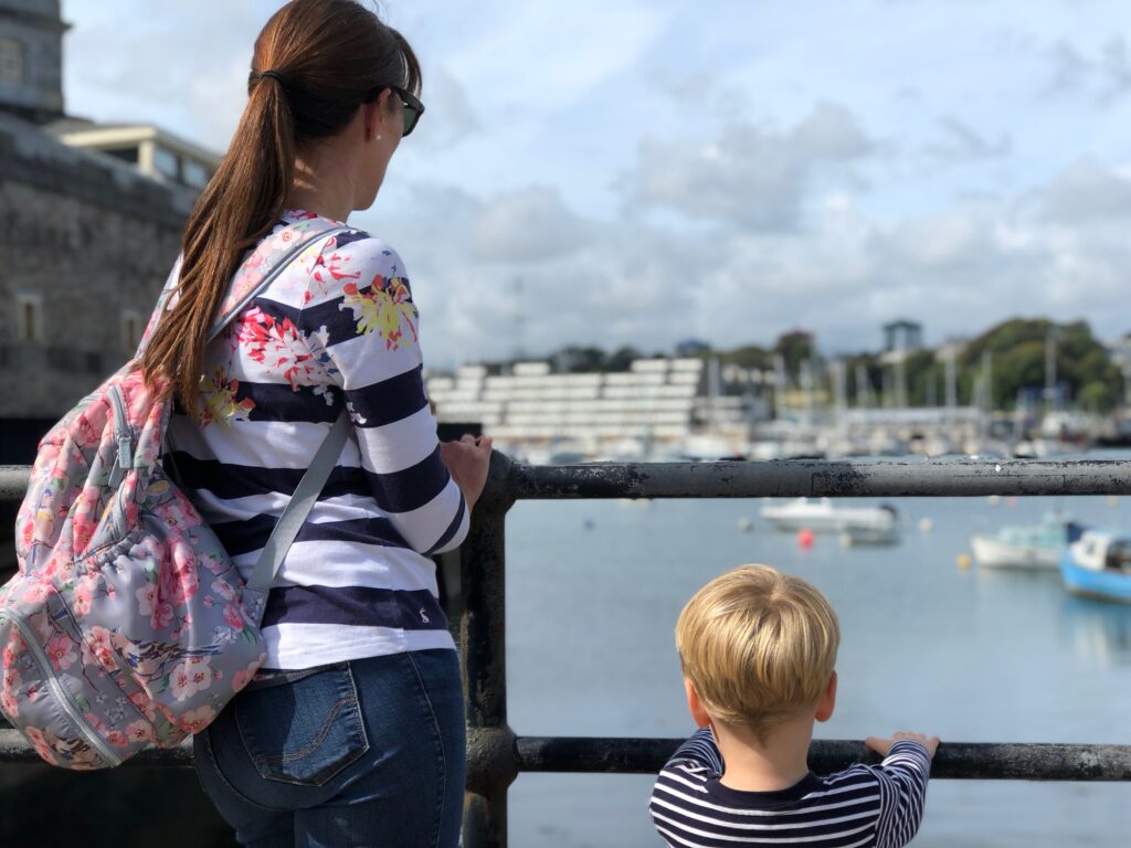 Woman with long brown ponytail and rucksack, wears a stripy top and jeans, and stands overlooking the sea and boats with a little boy wearing a stripy top.