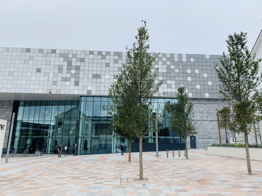 Modern building made up of tiny square pieces of glass on top of an old building to create the new museum The Box in Plymouth, with a tree and paving outside.