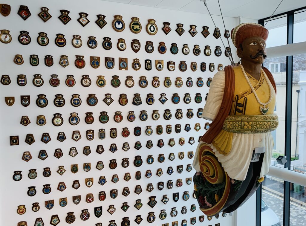 A giant wooden figureheads from naval ships preserved and restored hanging from the ceiling of The Box museum in Plymouth. The wall behind is covered with naval badges