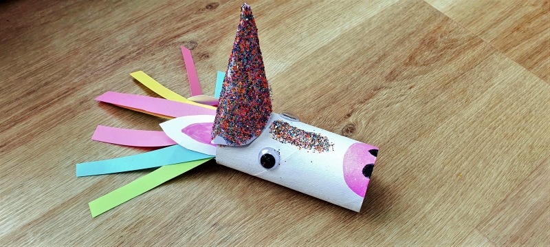 A toilet roll made into a unicorn with paint and glitter, with colourful pieces of paper as hair and a pink glittery horn, laid on a wooden table