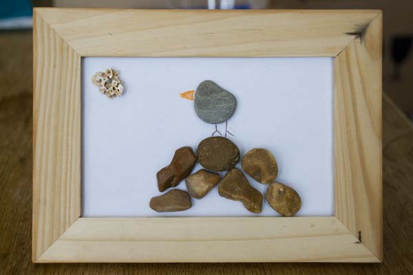 A wooden picture frame with a white sheet of paper inside with small flat pebbles stuck to it arranged in a picture to show a bird sat on a pile of rocks