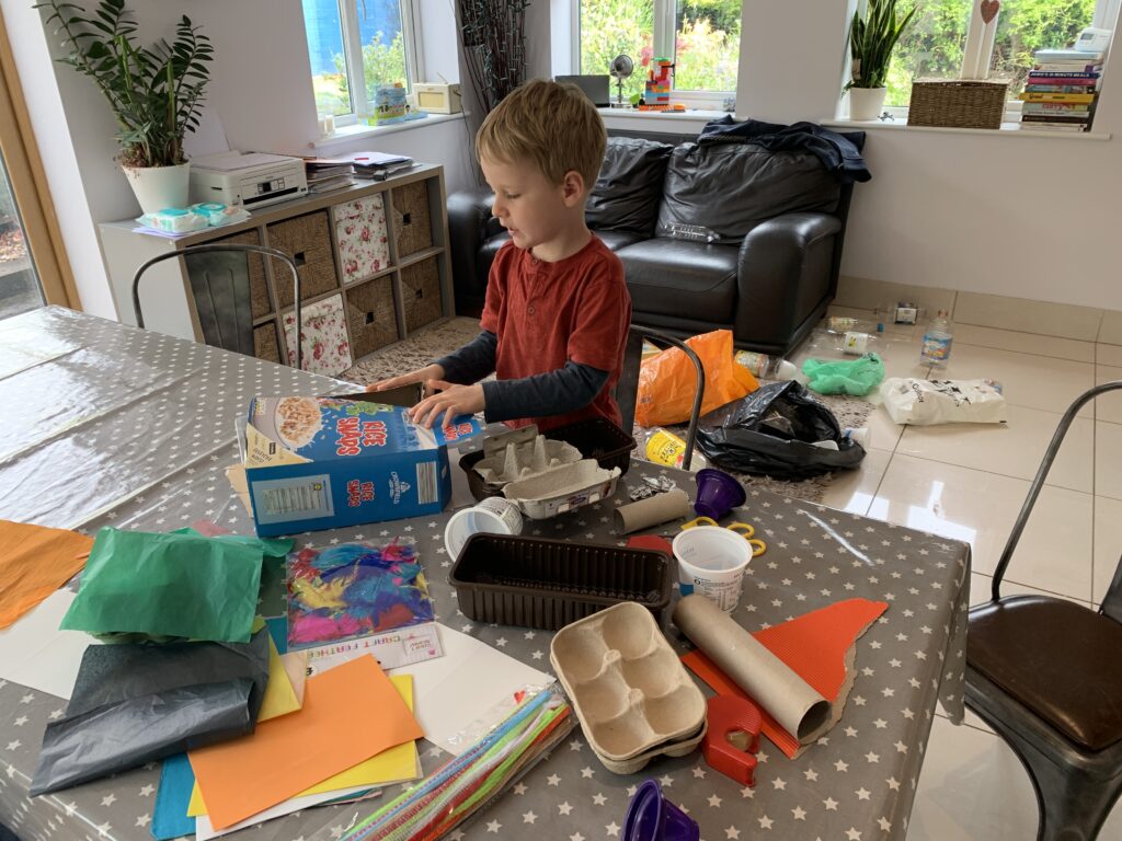 A five year old boy at a kitchen table with a settee in the background doing junk modelling. The table and floor behind him is strewn with items of recycling such as cereal boxes, plastic bottles, paper, egg boxes, yoghurt pots and kitchen roll holders.