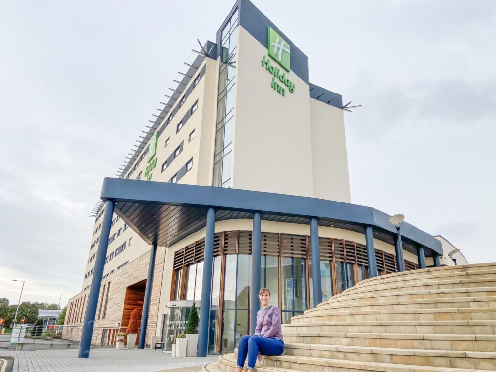 A woman in jeans and stripy jumper sat on some steps outside a tall Holiday Inn building