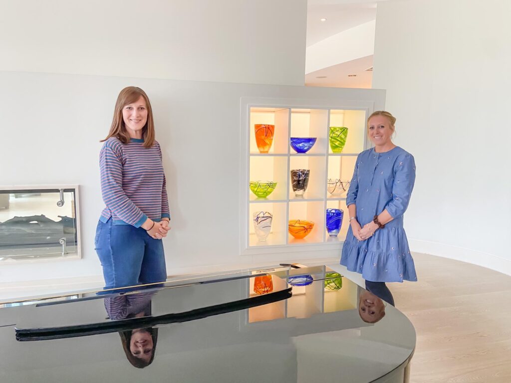 Two women standing next to a wall with a shelf of colourful vases and a grand piano in front