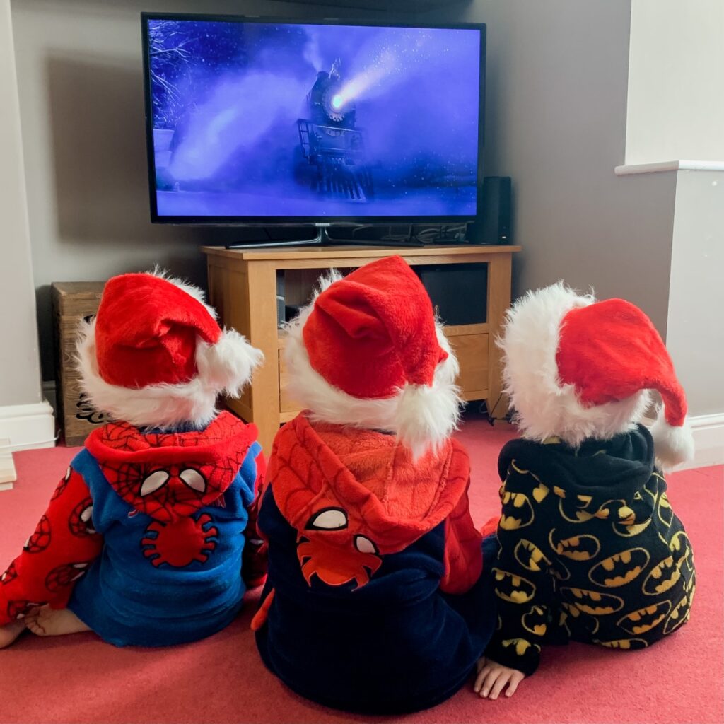 three boys wearing santa hats and dressing gowns sitting on a red carpet watching polar express on the television