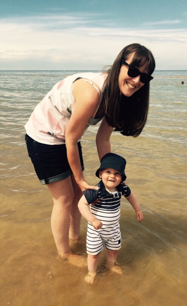 Long dark haired lady with sunglasses holding the arms of a little boy in a rompersuit paddling in the sea in France