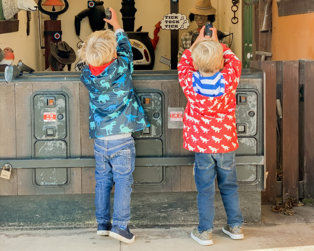 Three year old twin boys wearing a blue coat and red coat, and jeans stretch up to hold the guns on a shooting game at Pennywell Farm.
