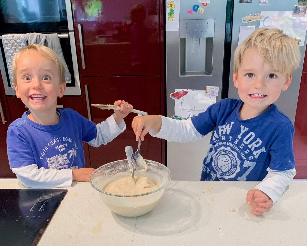 Three year old twin boys wearing blue t shirts stir the batter ready to bake a cake at a kitchen surface