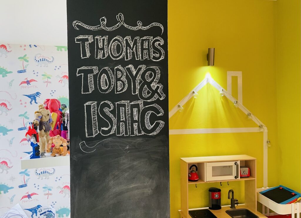 A strip of blackboard wall in a playroom with the names Thomas, Toby and Isaac written on it in white chalk