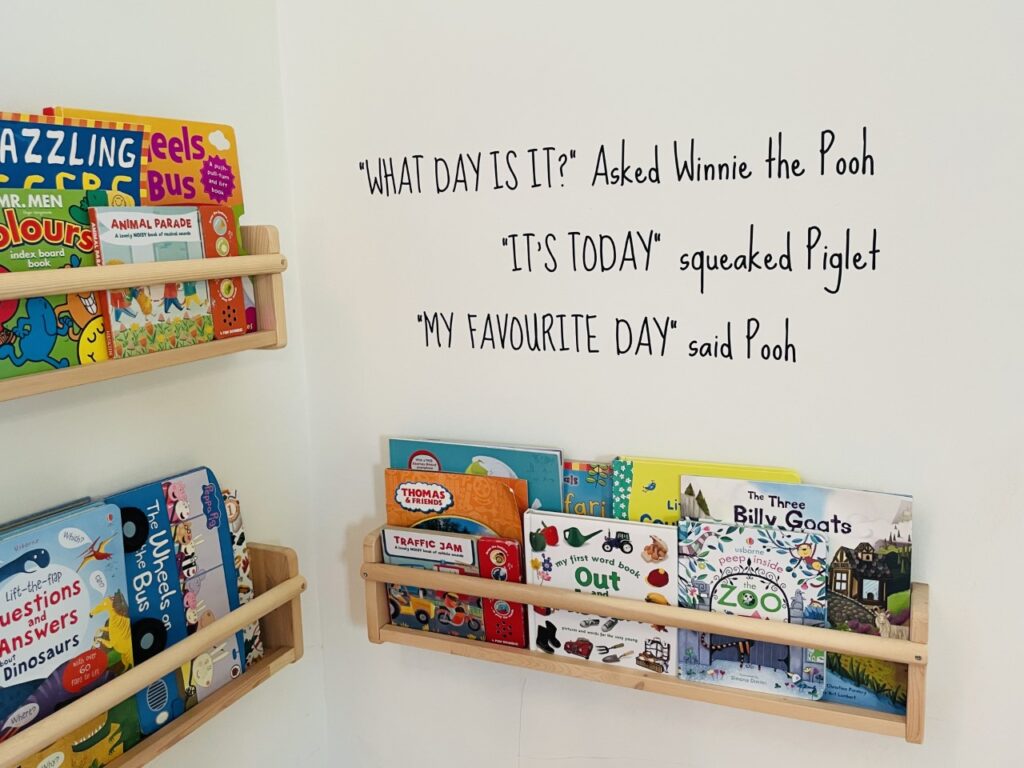 A wall with wooden bookshelves full of children's books plus a quote from Winnie the Pooh in black stickers on the wall