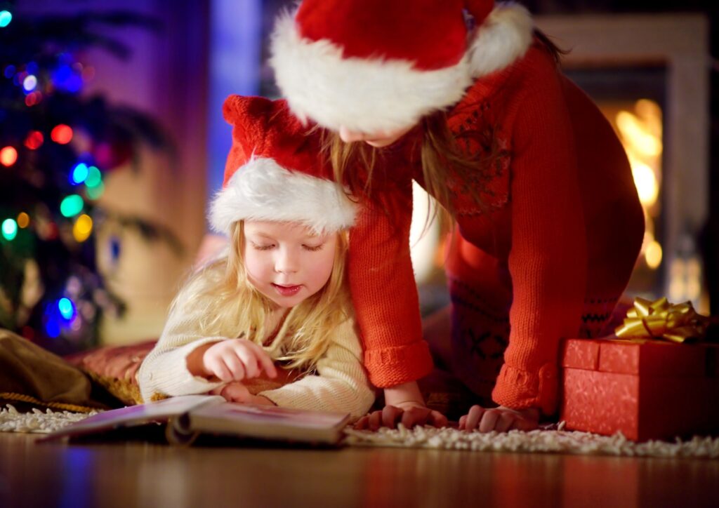 Little girl and mum in santa hats on the floor reading a book with a Christmas tree in the back ground