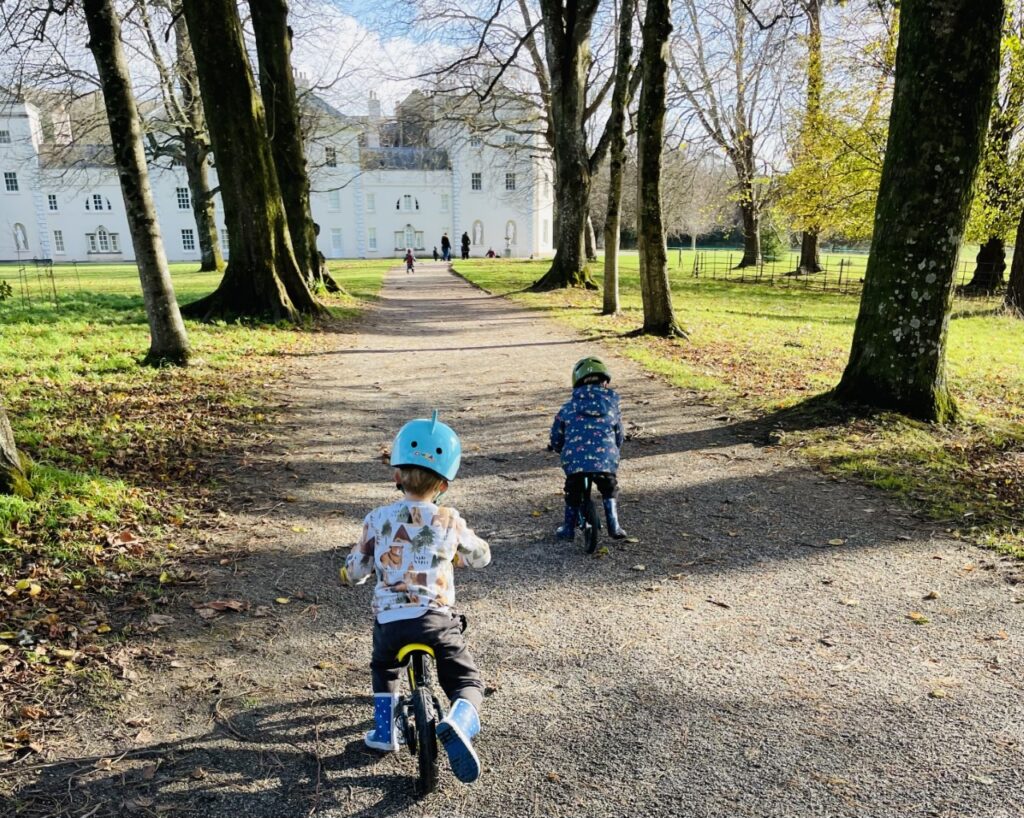 Two little boys on balance bikes riding along a tree lined avenue towards Saltram House as a Plymouth place to visit in Tier 3