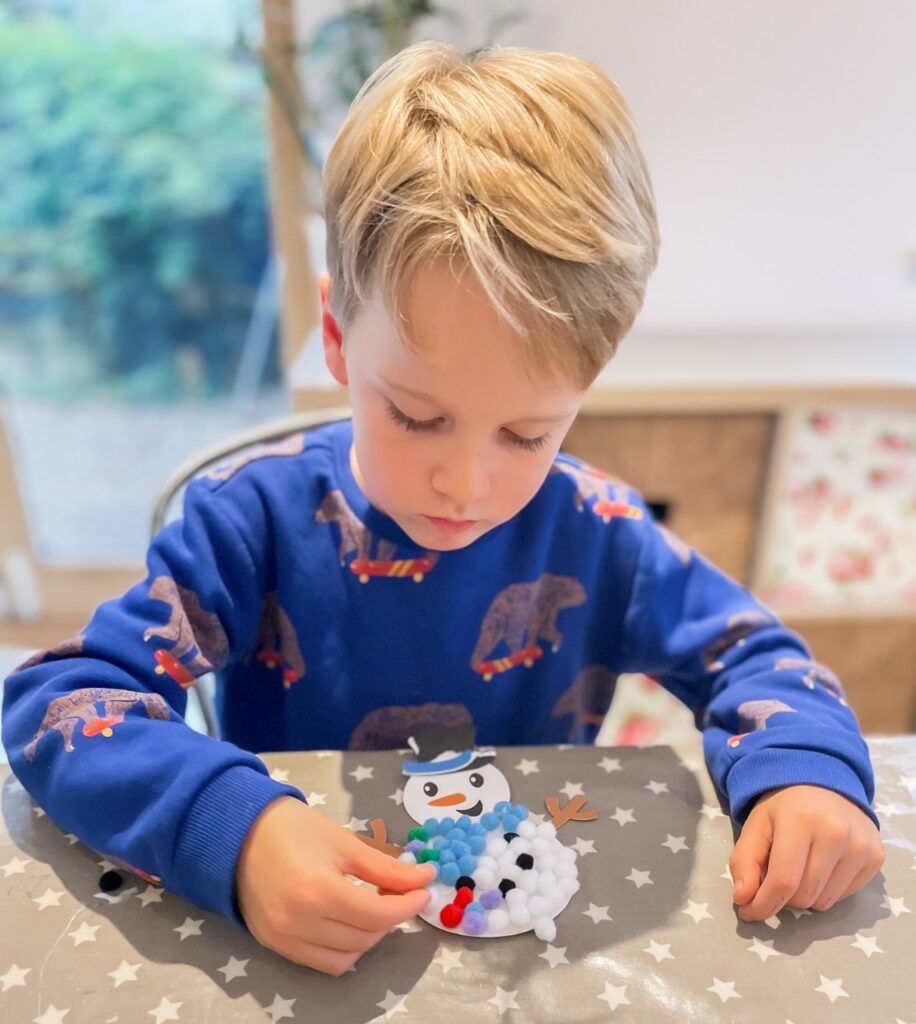 Five year old boy sat at a table doing christmas craft sticking pom poms onto a snowman