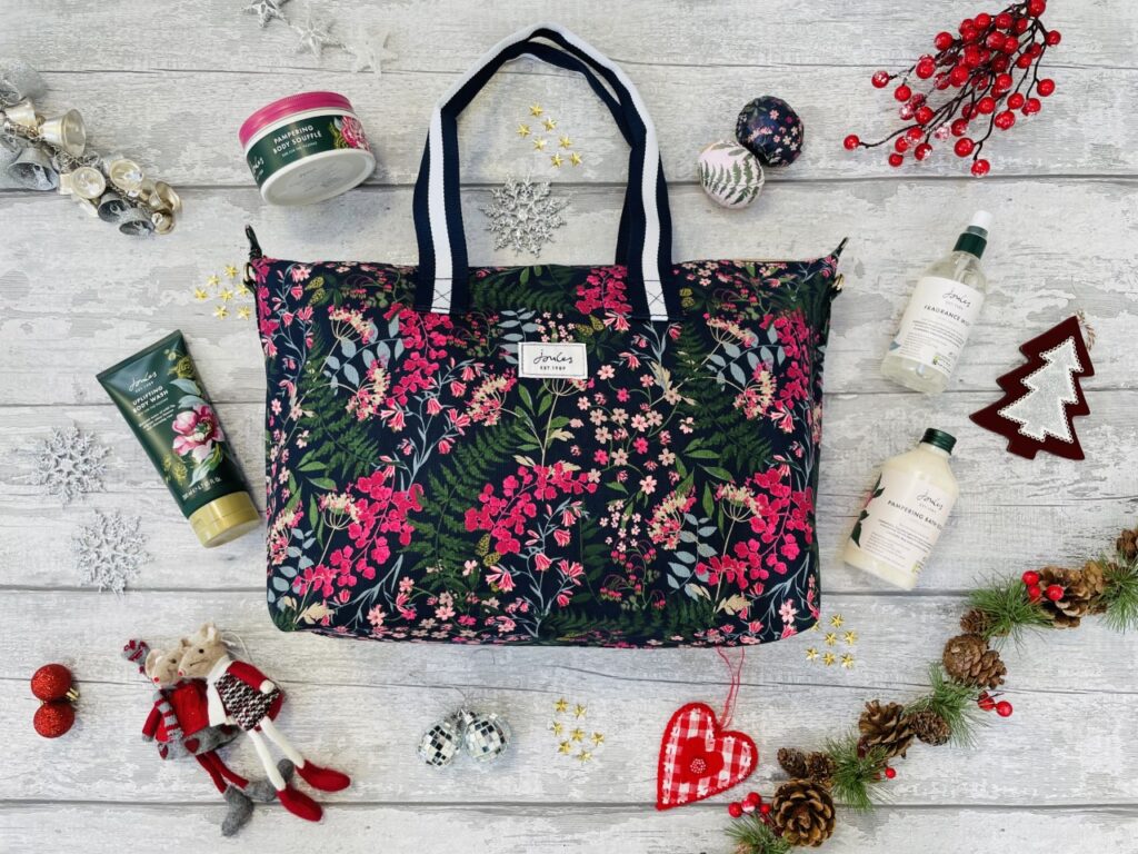 A pink and navy flowery joules bag with various bottles of pampering items on a grey background surrounded by christmas decorations