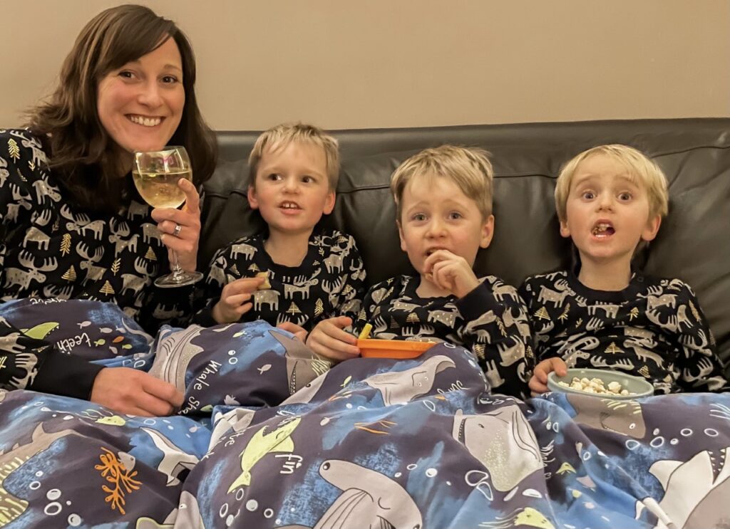 Mum and three boys in matching pyjamas keeping warm under a duvet on the sofa