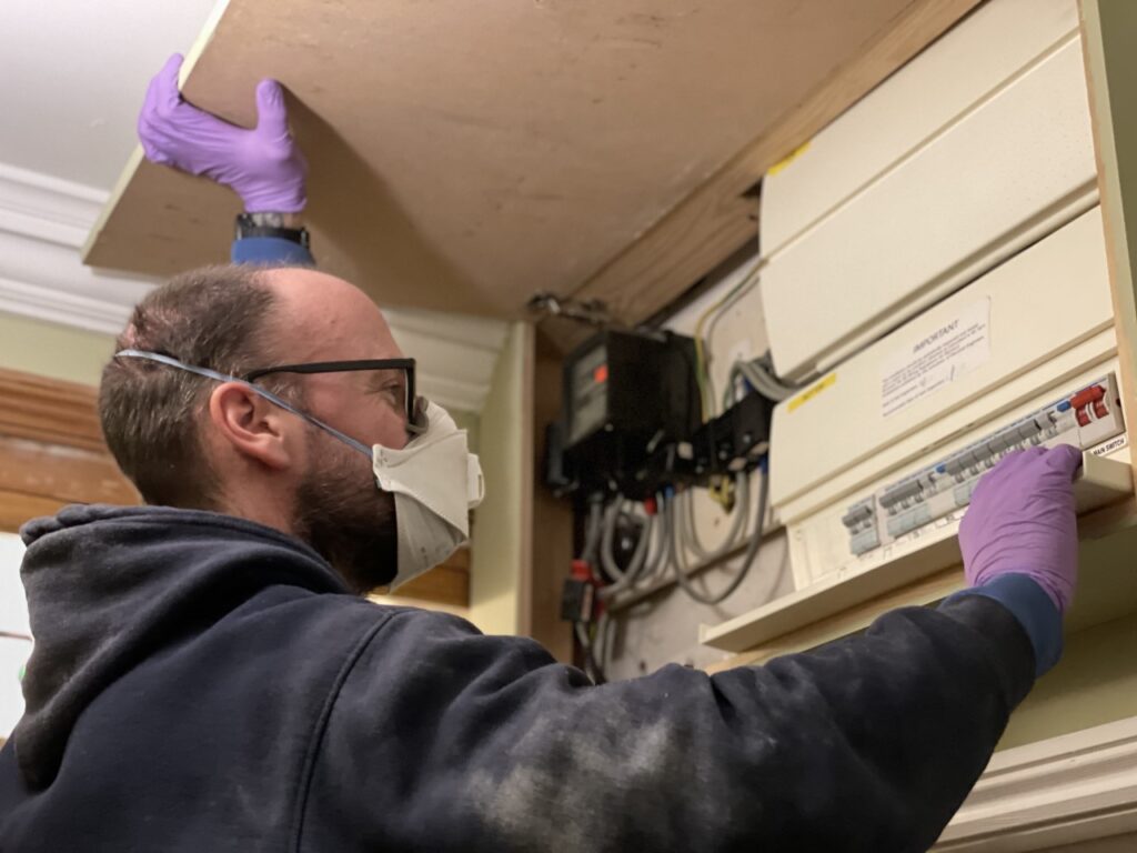 Electrician wearing a face mask and gloves checks a fuse box for a piece about tradespeople in your home during Covid