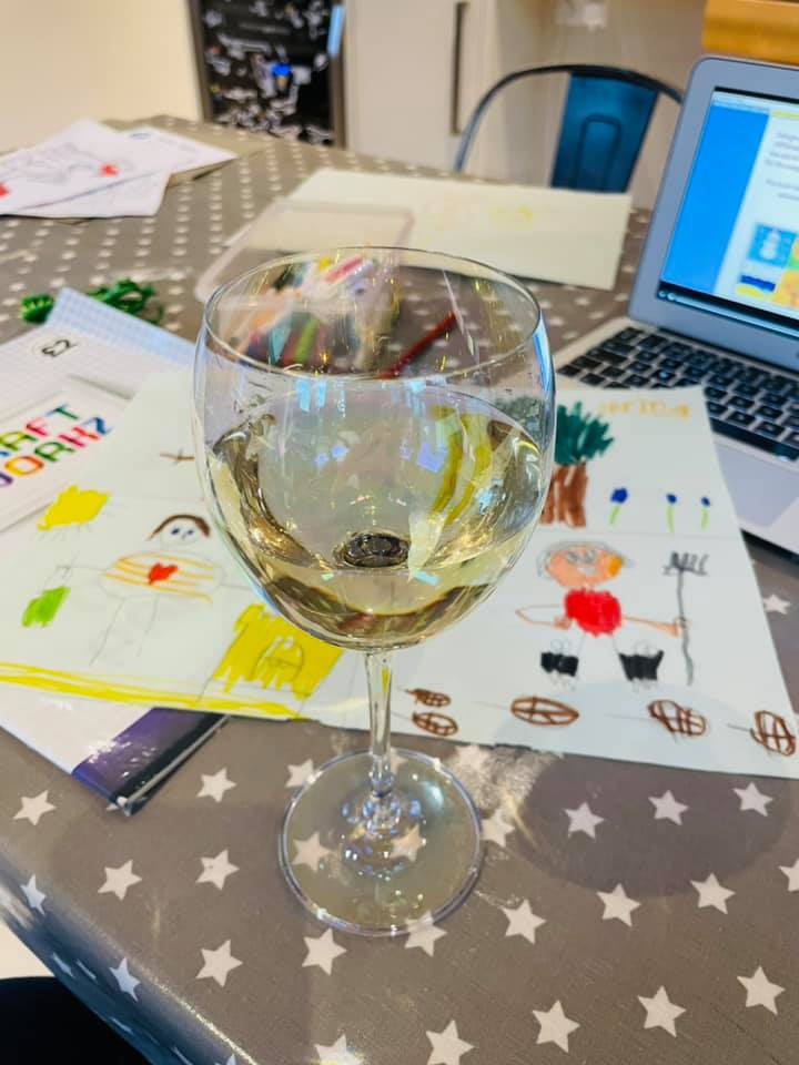 A glass of white wine in front of a laptop and children's colourings