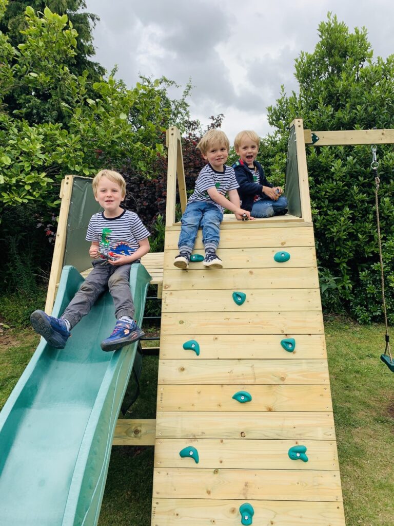 Three brothers sitting on the top of a wooden climbing wall and slide in their back garden