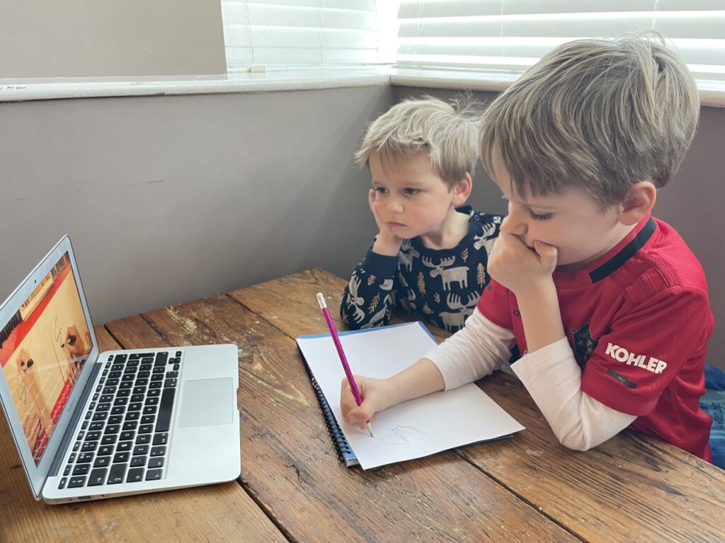 Five year old boy drawing on a bit of paper in front of a laptop on a table, with a three year old boy watching on