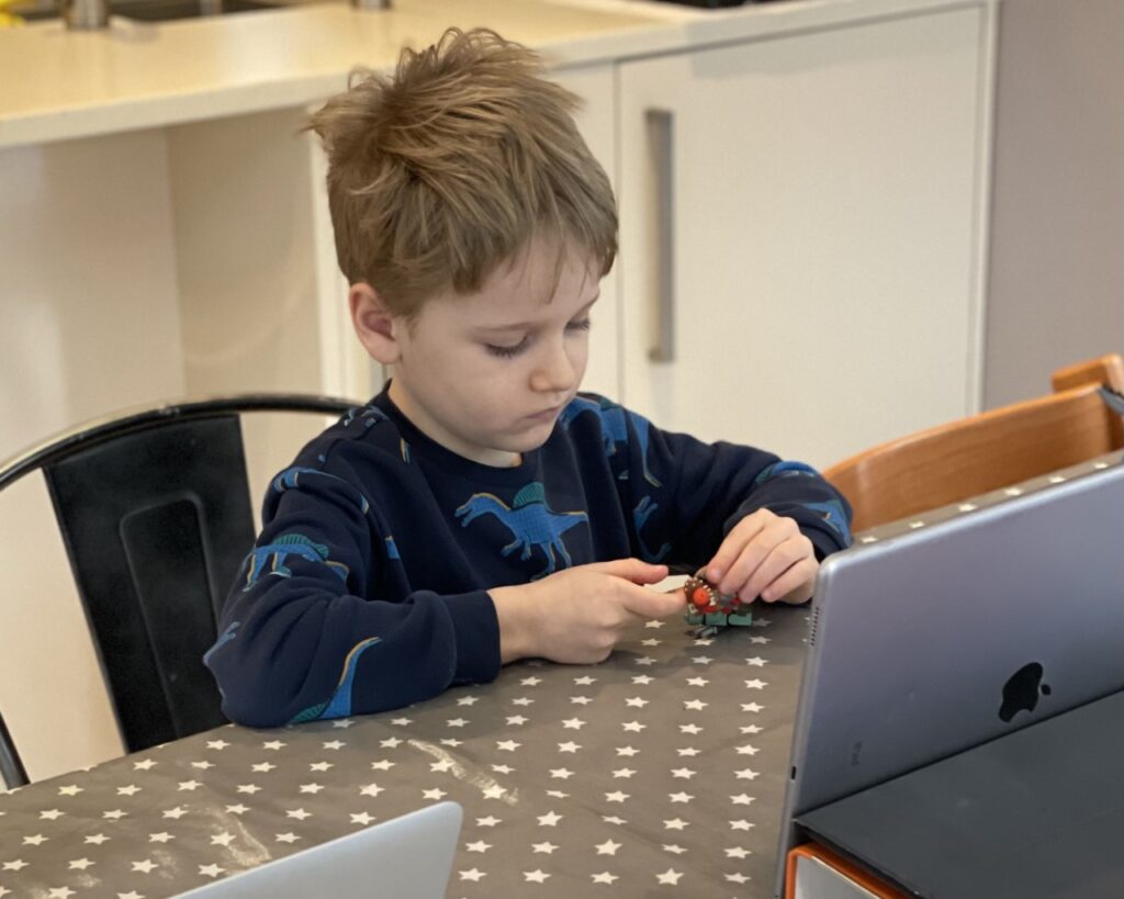 Five year old boy sitting at a table playing with a lego mini figure during a homeschooling video