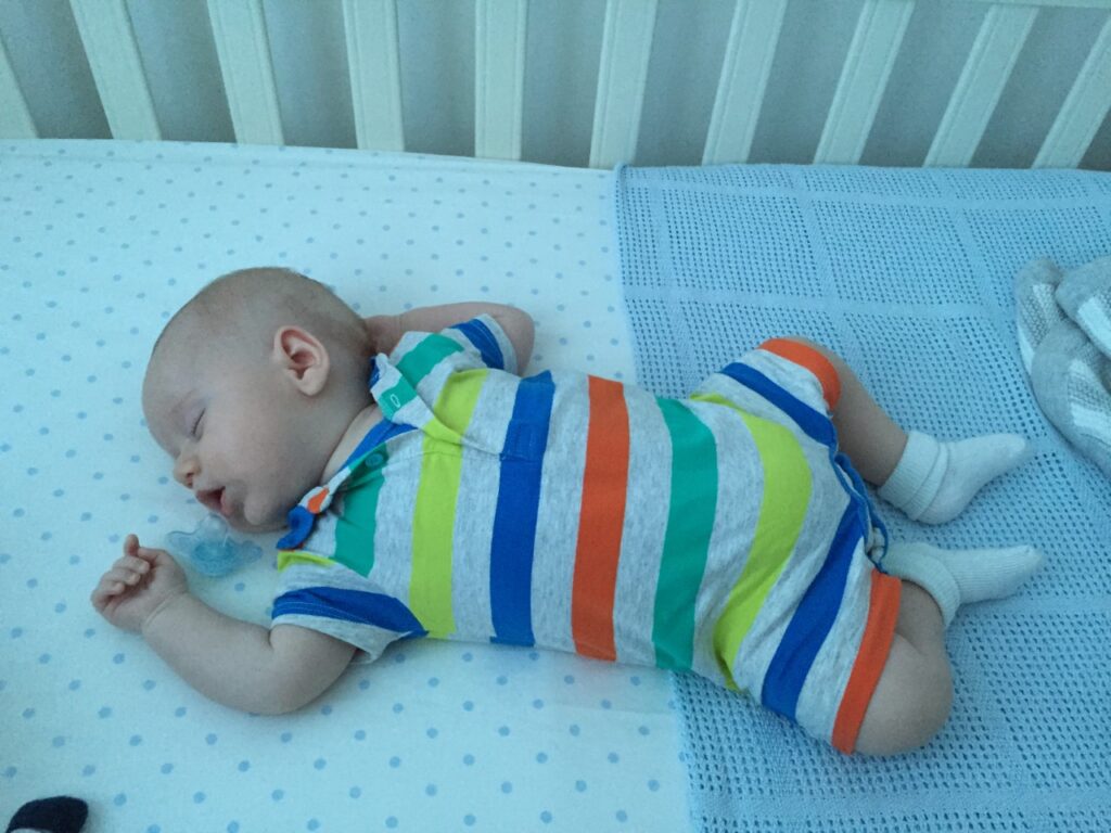 Baby wearing a stripy romper asleep in his cot