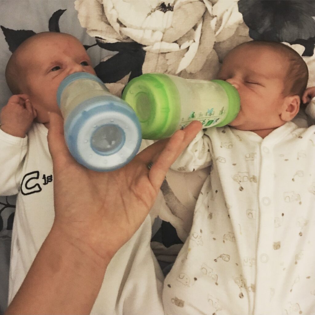 Newborn twins being bottle fed on a bed. Mum's hand holding both bottles.