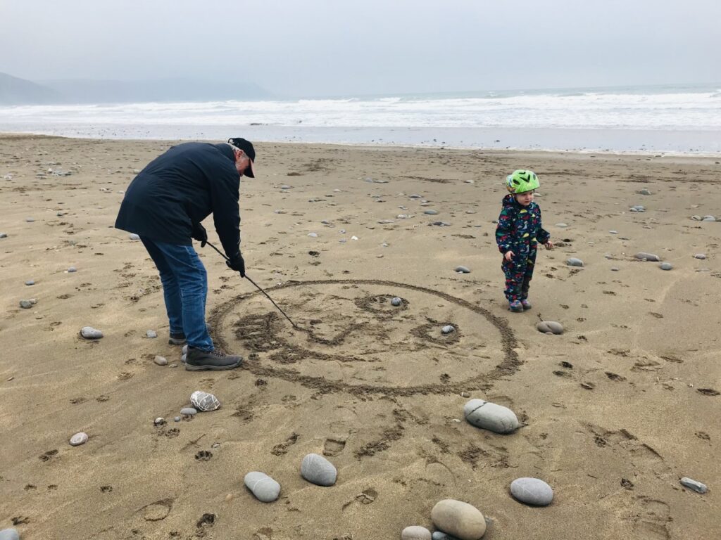 A grandad and two year old draw a face in the sand on the beach