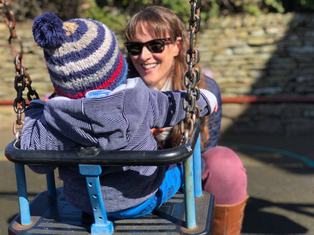 Mum with dark hair and sunglasses crouched down in front of a baby in a swing