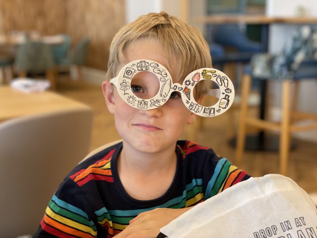 Six year old blonde boy wears funny oversized wooden glasses at a restaurant table