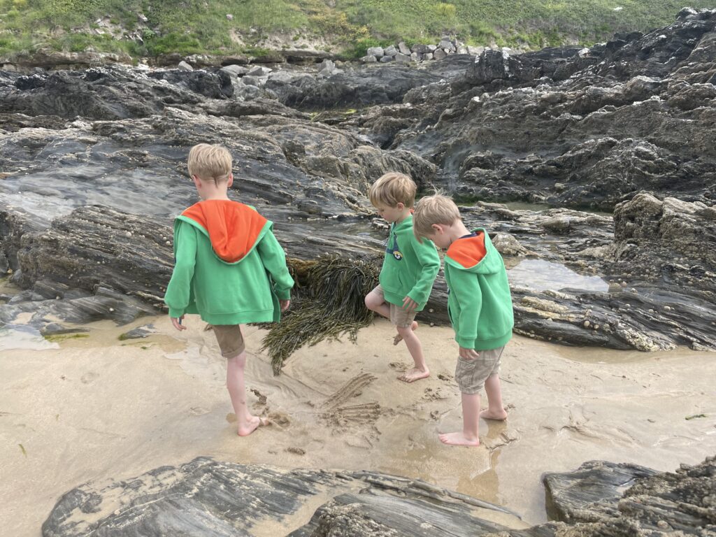 Three young boys in shorts and green hoodies play in rockpools on Fistral Beach in Cornwall