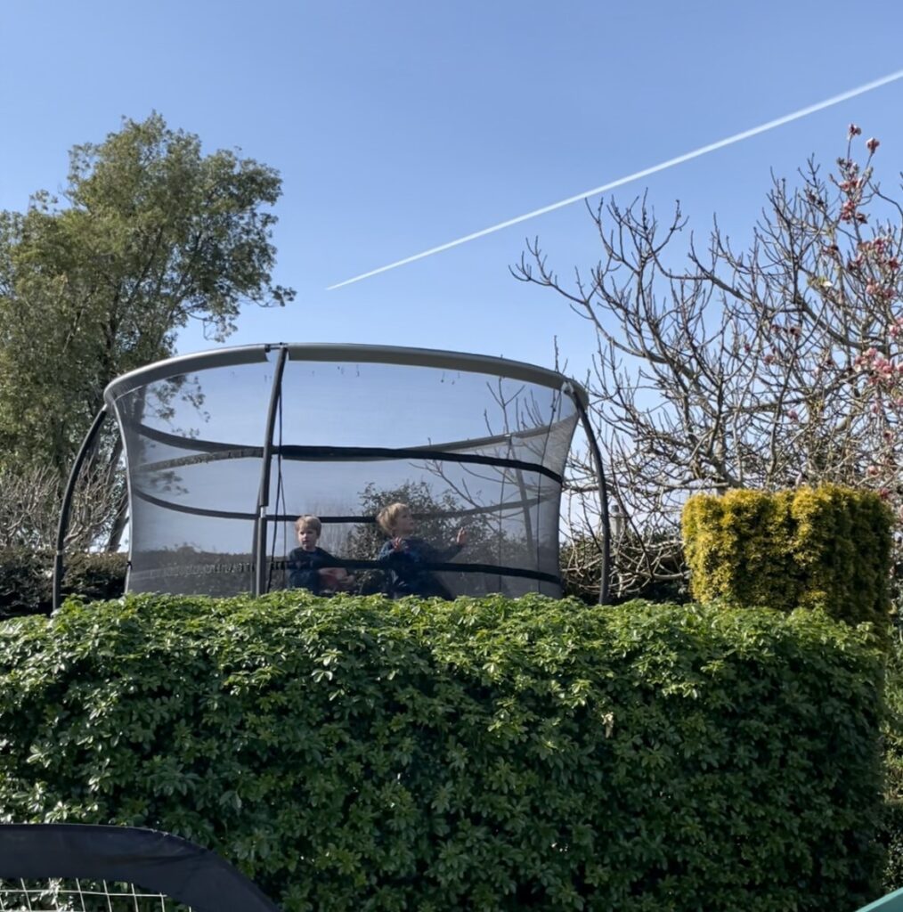 Twin three year olds bounce on a large trampoline with a bright blue sky in the background and an aeroplane trail