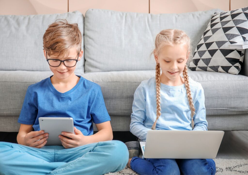 Blonde boy and girl wearing blue sitting on the floor against a settee watching a laptop and tablet to learn a second language as well as do coding lessons