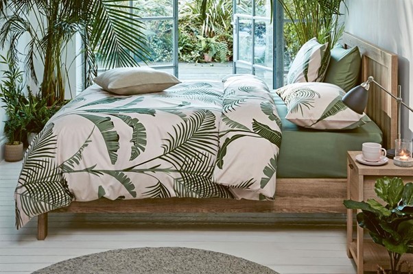 A bed with plants on the duvet cover and green sheets, with plants in the corner of the room in a Tropics style decor