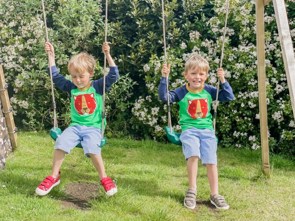 Twin boys wearing shorts and long sleeved tops sit on a double swing set in the garden