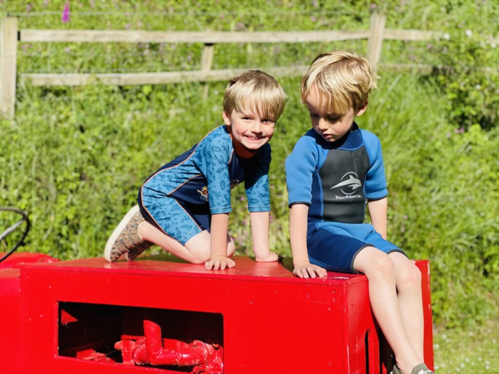 Four year old twin boys in wetsuits sat on the top of a tractor at a park before swimming