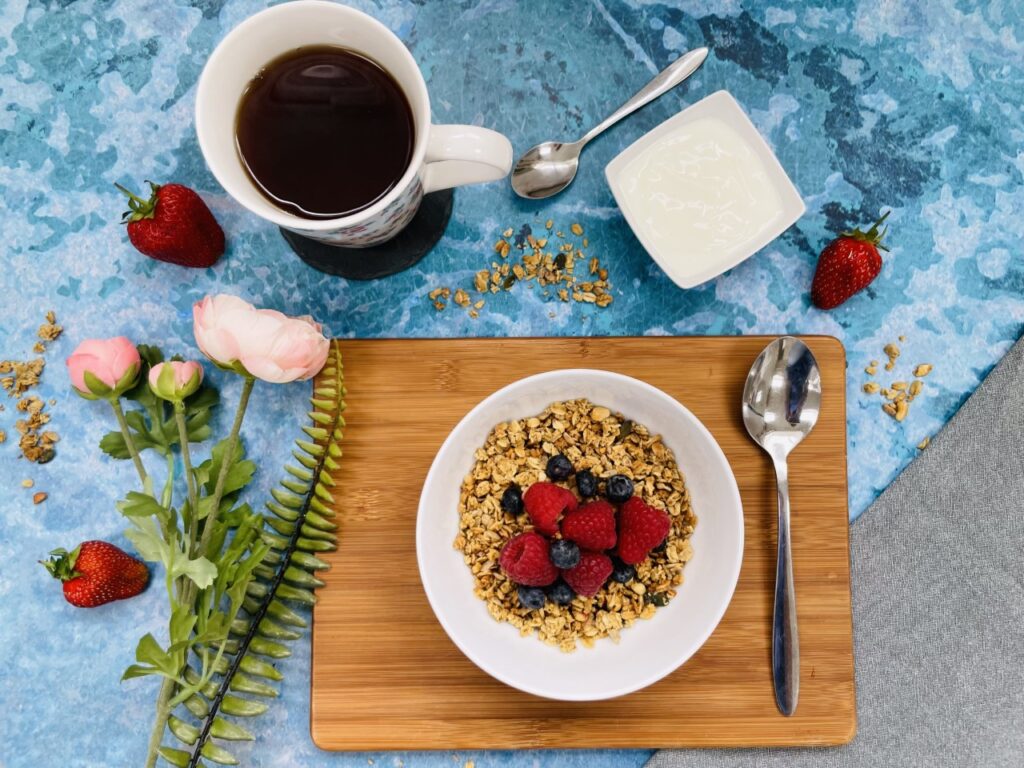 A blue flatlay of a bowl of granola containing good bacteria, a black coffee and some flowers