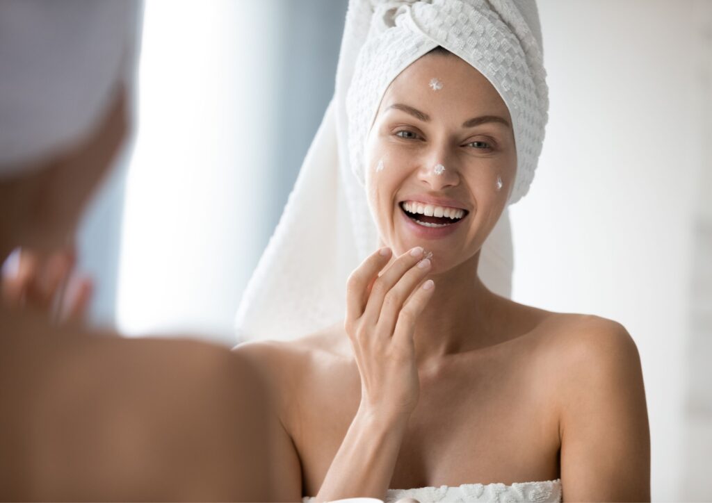 Woman with hair in towel puts cream on her face as part of a skincare routine great for health and beauty essentials