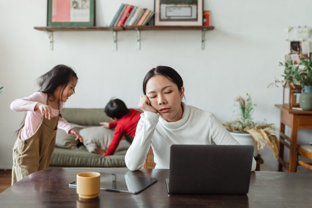 A woman on a laptop at a table working from home with kids with the kids on the settee in the background